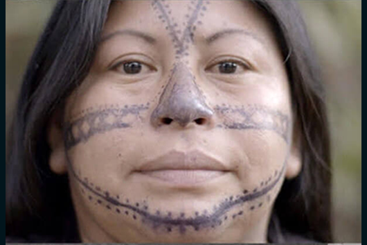 still from Amazonia; an indigenous woman with face tattoos gazes into the camera